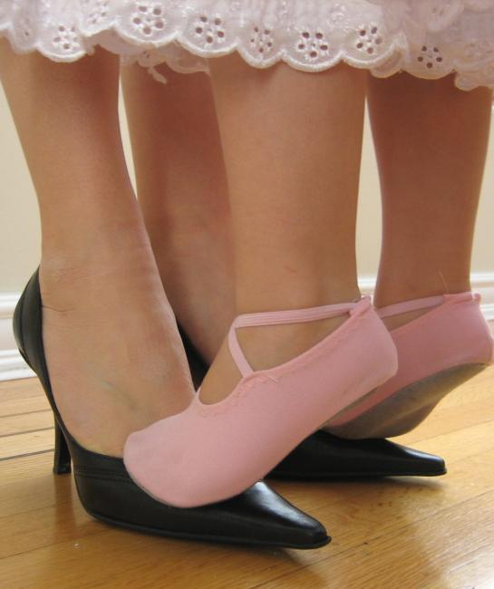 A little girl in pink ballet slippers stands on her mother's feet