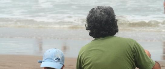 father and son sitting on the beach together watching waves and surfers