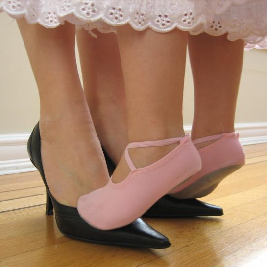 A little girl in pink ballet slippers stands on her mother's feet
