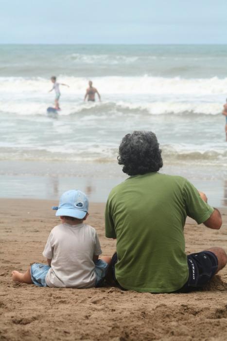 father and son sitting on the beach together watching waves and surfers