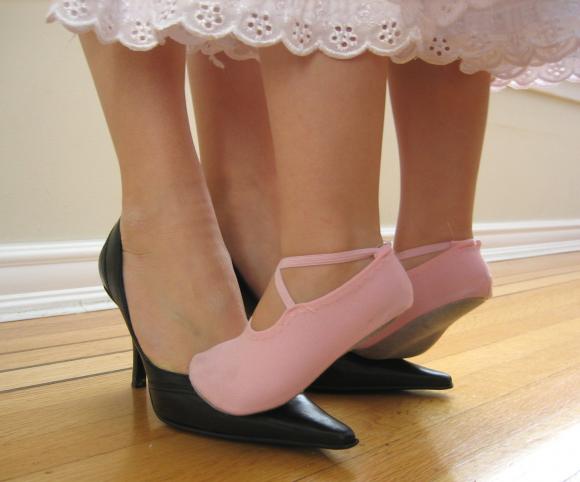 A little girl in pink ballet slippers stands on her mother's feet